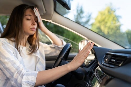 A/C Repair & Services in Canton, CT | Collinsville Auto Repair. Image of a woman driver having problem with a non-working conditioner, her hand checking flow of cold air, wiping sweat from her forehead with tissue.