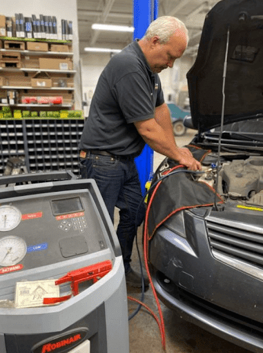 Car AC Blowing Hot Air | Collinsville Auto Repair in Canton, CT. Image of a male mechanic refilling the refrigerant of a car with an AC gas recharging machine. The refilling takes place in an auto repair shop.